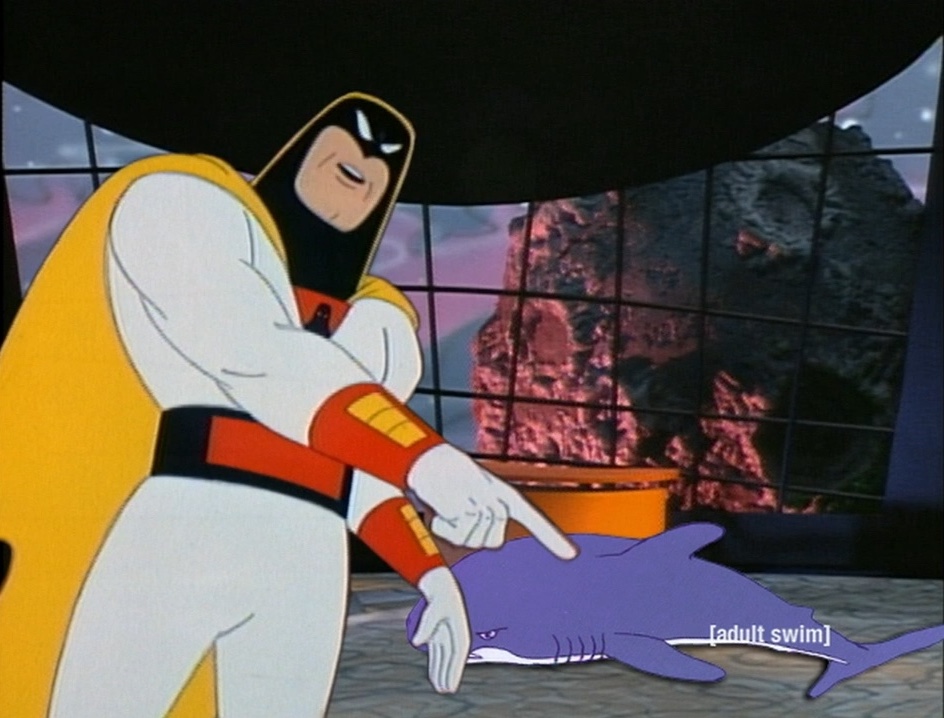 A picture of the Space Ghost Coast-to-Coast talk show desk. Space Ghost is standing in front of it, pointing at a shark lying on the ground